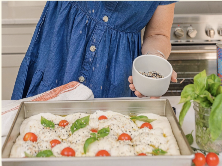 Events A person in a denim dress sprinkles seasoning onto focaccia dough with tomatoes and basil in Williamson County, Tennessee.