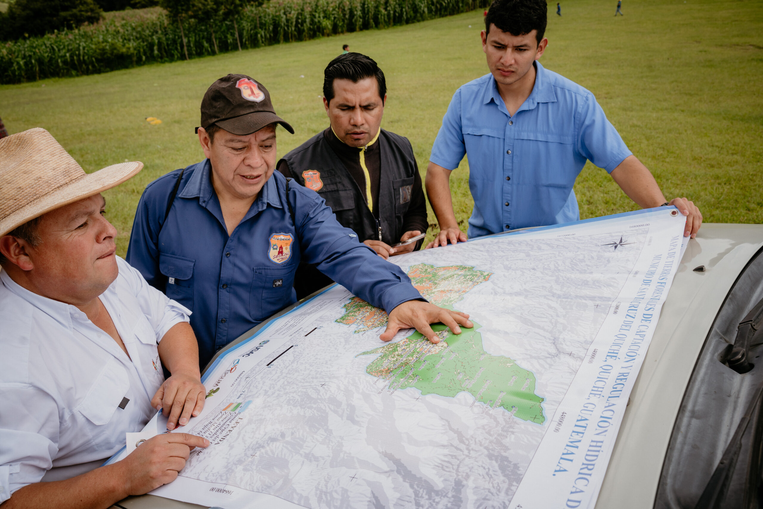 A group of four men view a map on the hood of a car with fields behind them