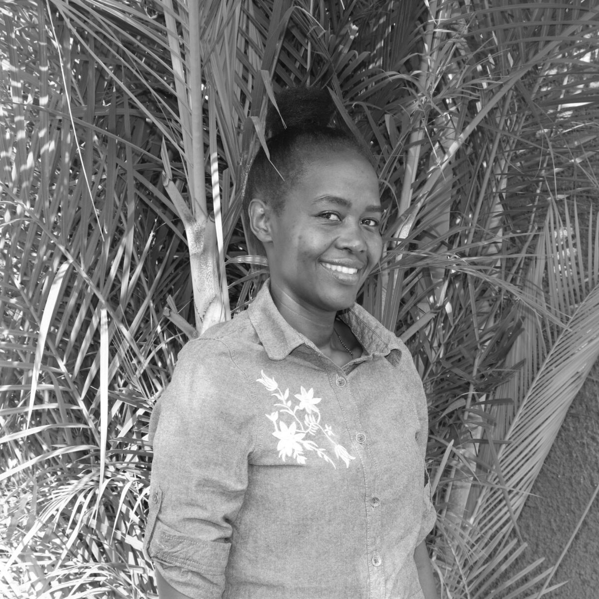 portrait black and white of Grace Kanweri, author of this article and senior program officer at Water For People Uganda. She is smiling at the camera against a palm tree background. 