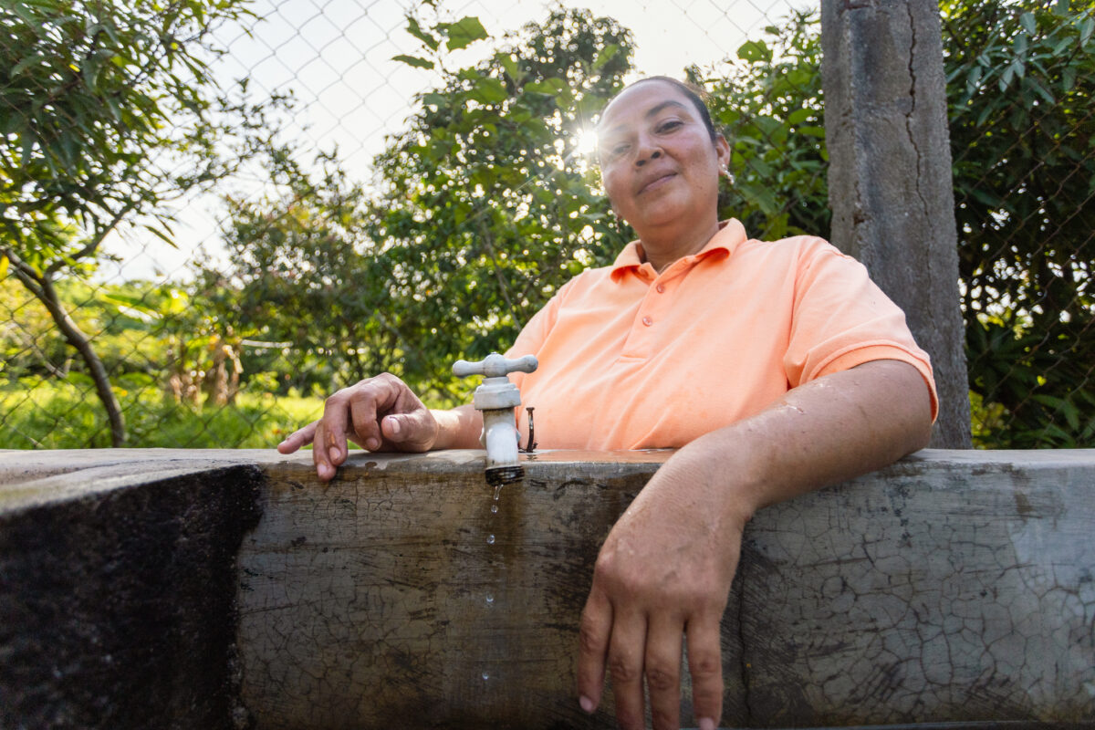 Carmen, A community water leader stands beside a concrete water tank with a faucet, demonstrating women-led clean water access in La Tigra.