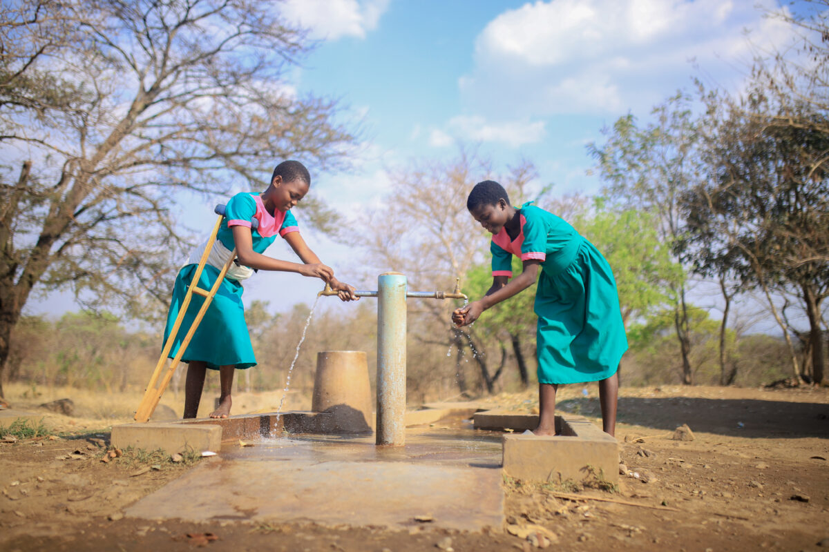 Two schoolgirls in green uniforms collect water from a hand-operated pump at a rural school, with one girl using crutches while pumping water.
