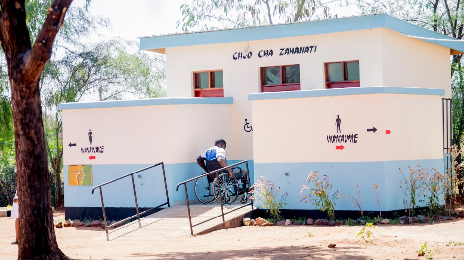 Person using a wheelchair accesses an inclusive latrine at Nghambi health clinic via a concrete ramp with handrails.