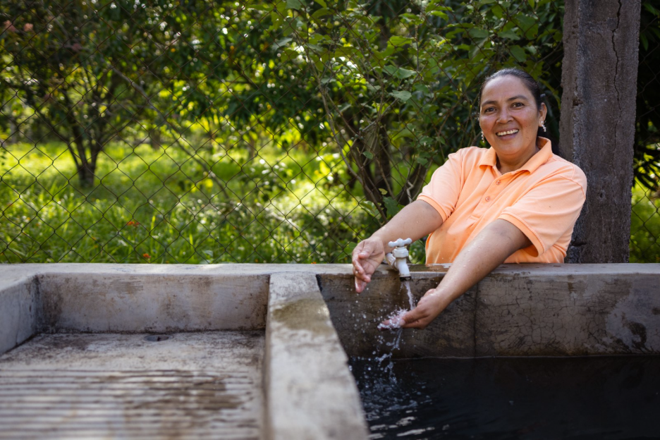 Carmen washes hands under a running faucet at a concrete water tank, showing clean water flowing reliably in La Tigra, Honduras, with green trees and fencing in the background.