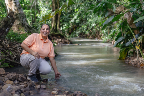 Carmen crouches beside a shallow stream in a forested area, touching the flowing water that serves as a vital source for the community.