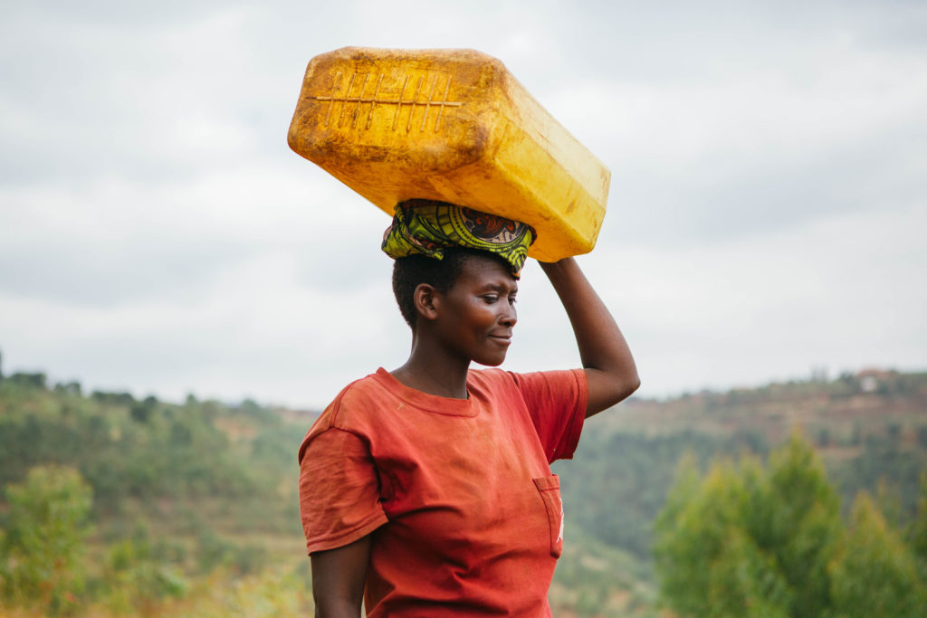 Woman in Rulindo in red with mountainous background, carrying water container.