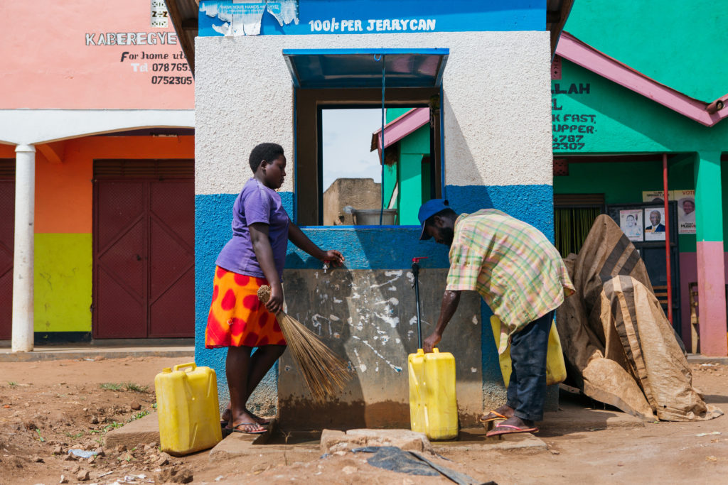A man fills a jerry can at a brightly colored water kiosk, while the kiosk attendant stands nearby